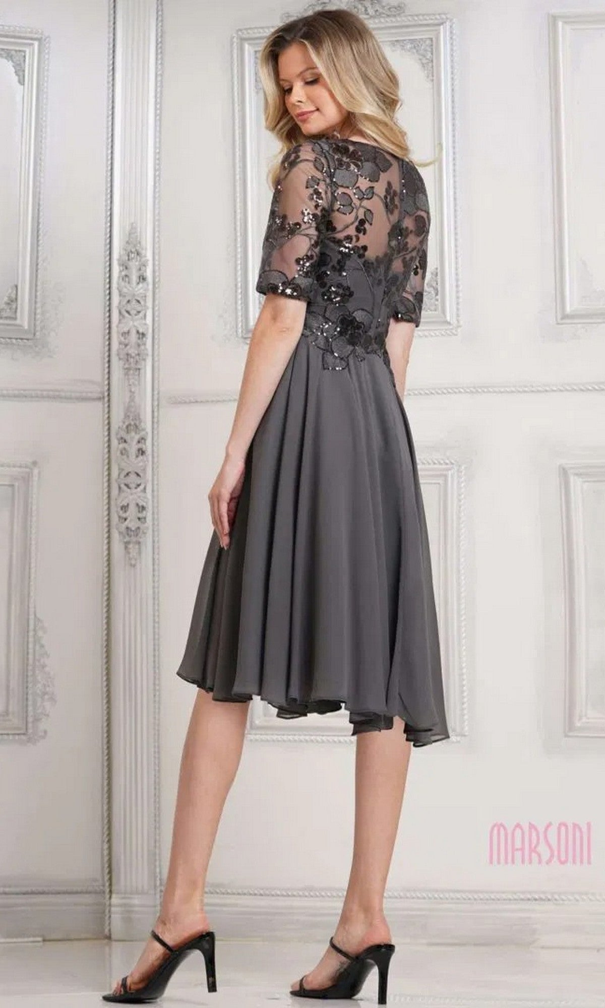 A woman wears the Marsoni M286S semi-formal knee-length dress with floral sequin lace on the bodice and sleeves, an elegant A-line chiffon skirt, and black heeled sandals while standing in an ornate room.